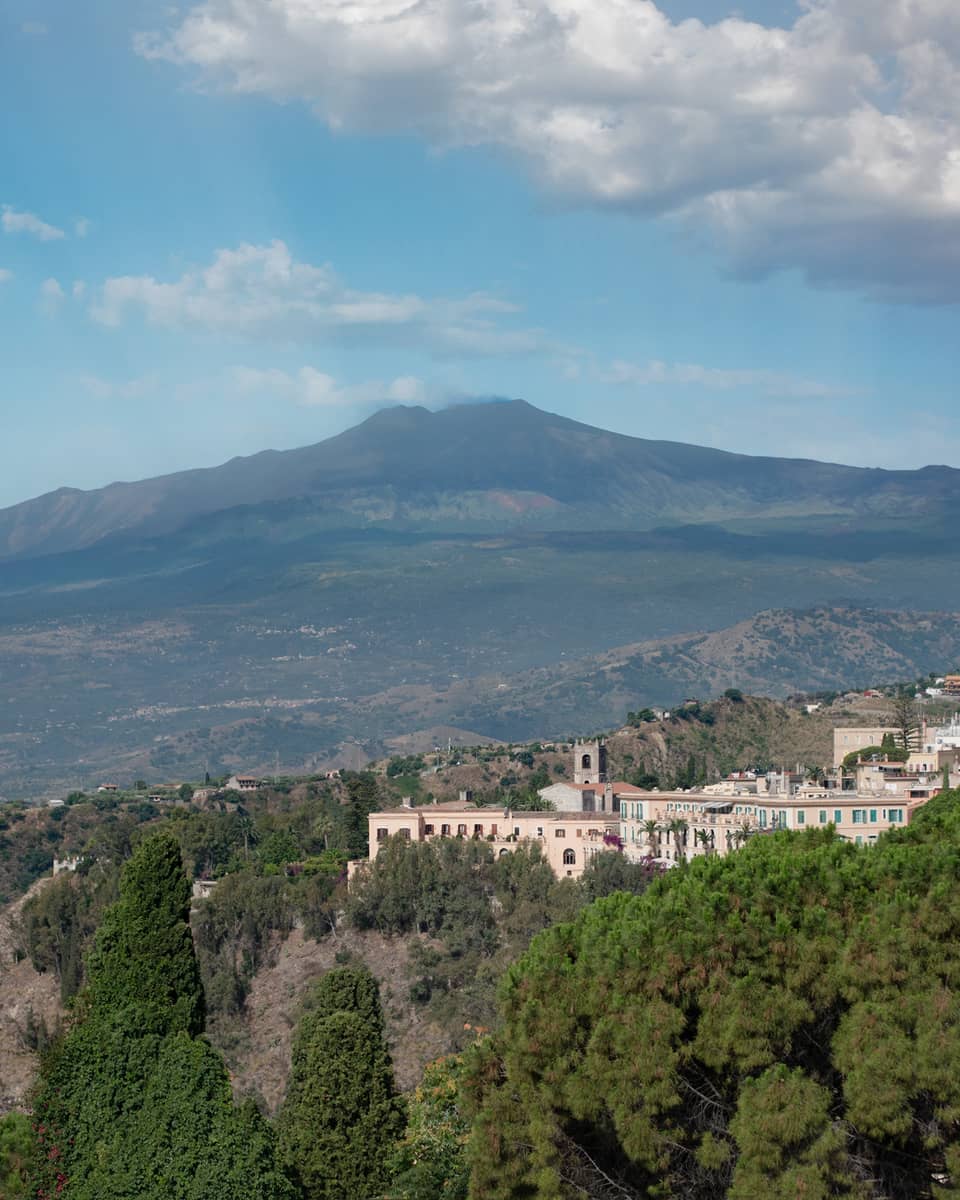 Hillside buildings surrounded by trees, Mount Etna and blue sky in backdrop