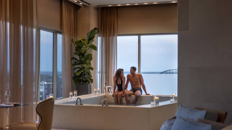 A couple enjoying a candlelit hot tub in a luxury hotel spa, with elegant seating, soft lighting and large windows offering a view of a distant bridge