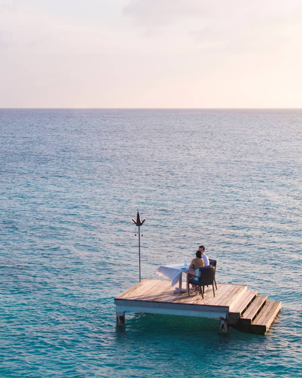 Man, woman dine at private table on small floating wood platform in middle of blue lagoon