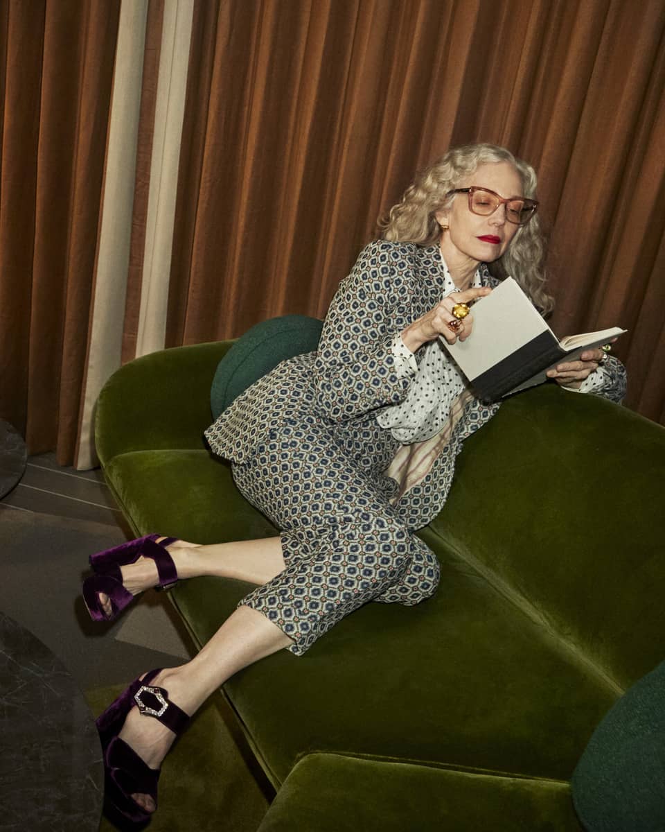 Stylish person with long gray hair wearing a patterned suit and purple heels sitting on a green sofa while reading a book