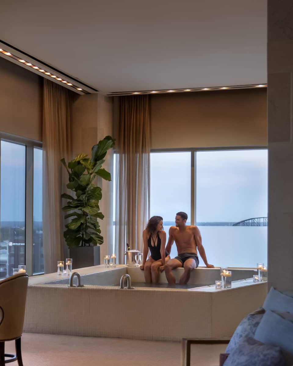 A couple enjoying a candlelit hot tub in a luxury hotel spa, with elegant seating, soft lighting and large windows offering a view of a distant bridge