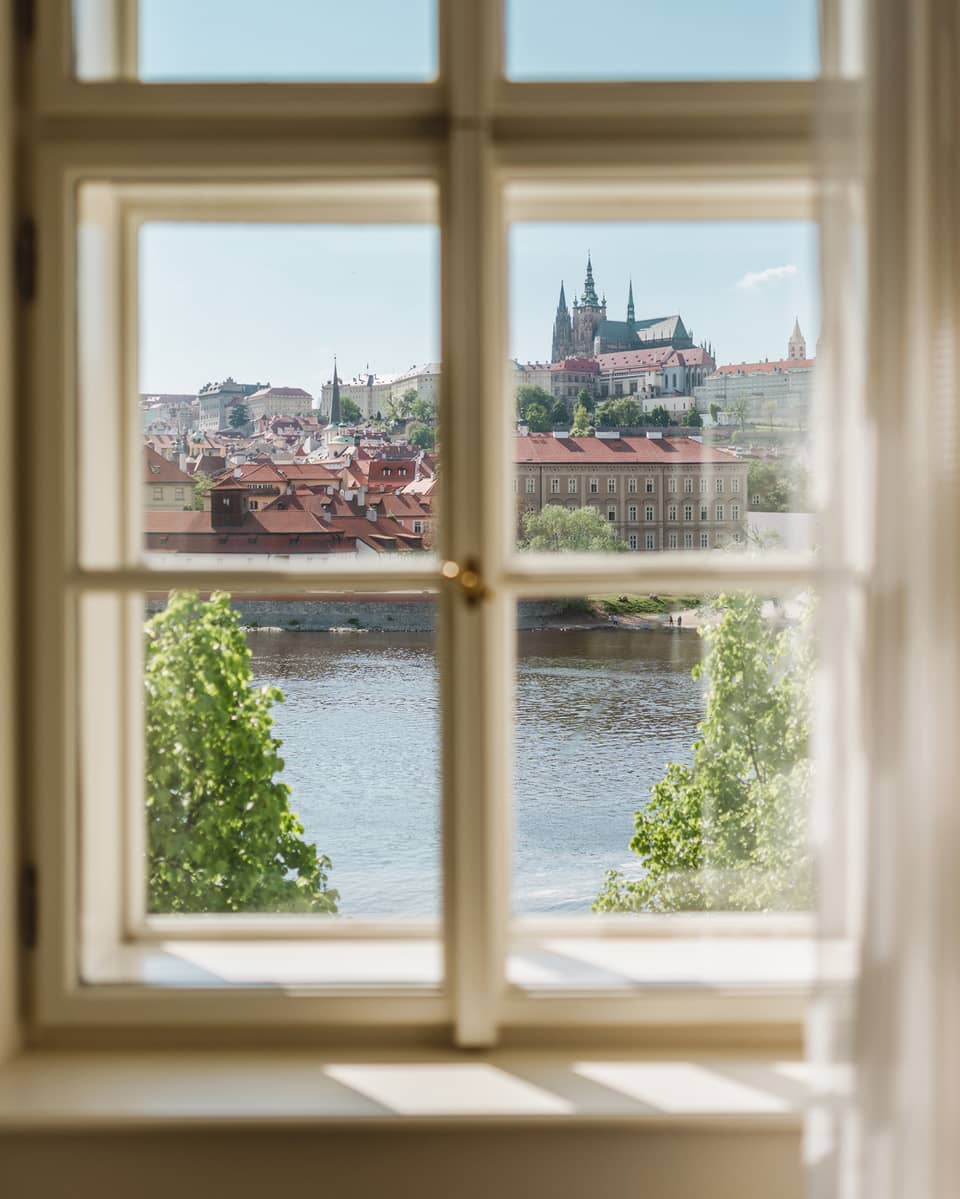 View through a window of a historic city with red rooftops and a cathedral in the distance, overlooking a river and green trees under a clear blue sky.