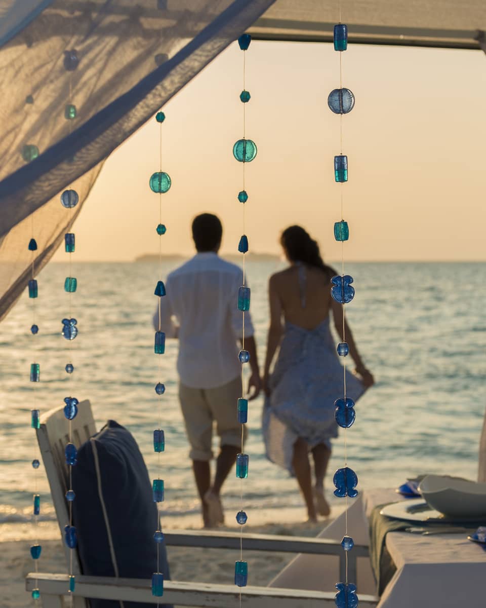 Sheer curtains blow in breeze, small blue beads hang from string, woman and man hold hands on beach in background