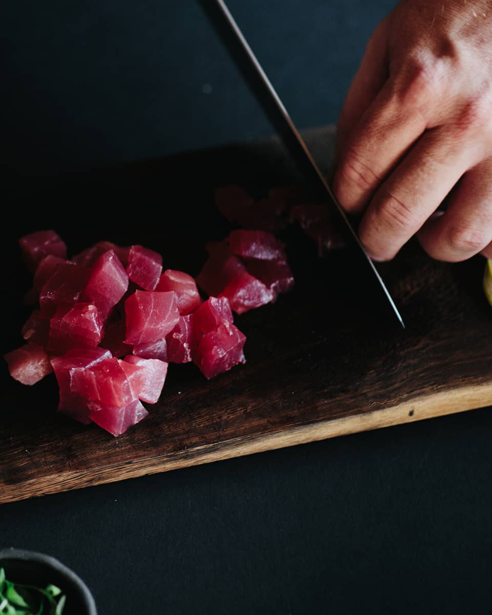 Man slicing raw tuna into cubes with knife on wood cutting board next to bowl of orange rock salt