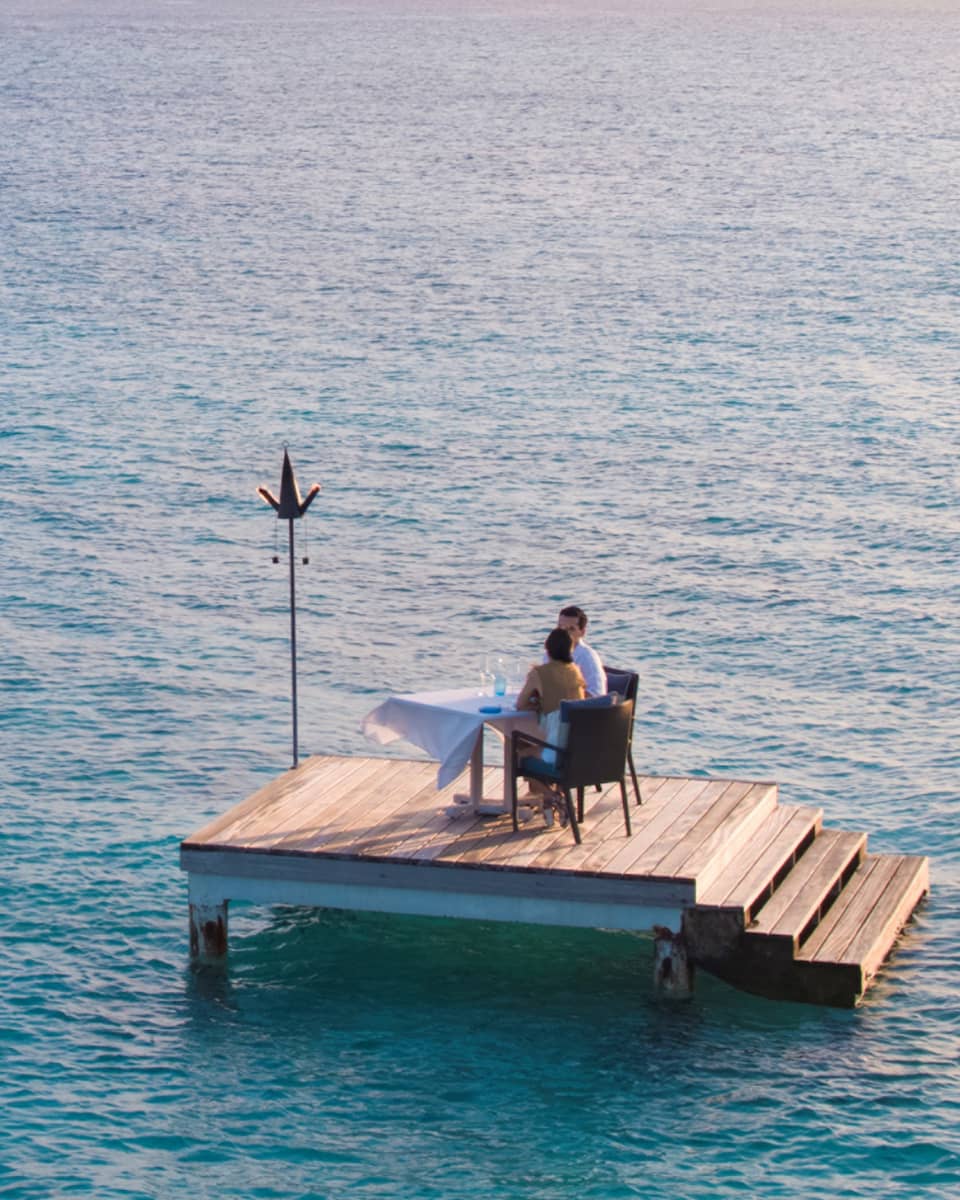 Man, woman dine at private table on small floating wood platform in middle of blue lagoon