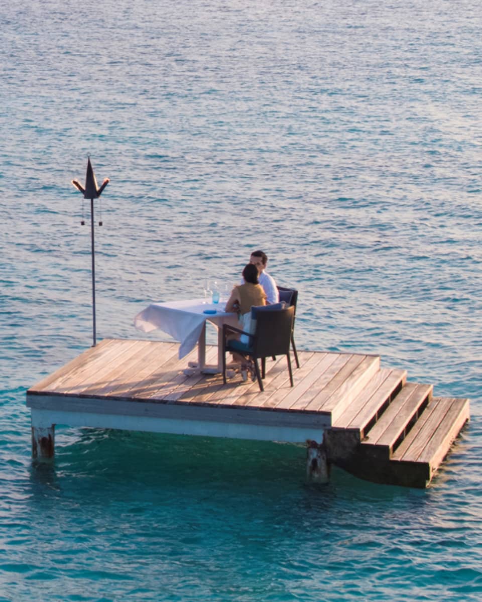 Man, woman dine at private table on small floating wood platform in middle of blue lagoon