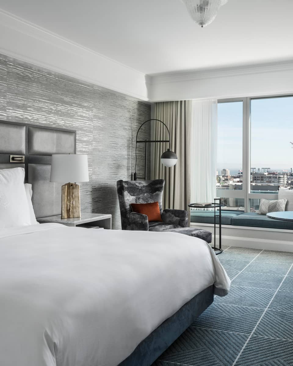 Modern hotel room with king bed, table and two upholstered grey chairs, windows overlooking San Francisco