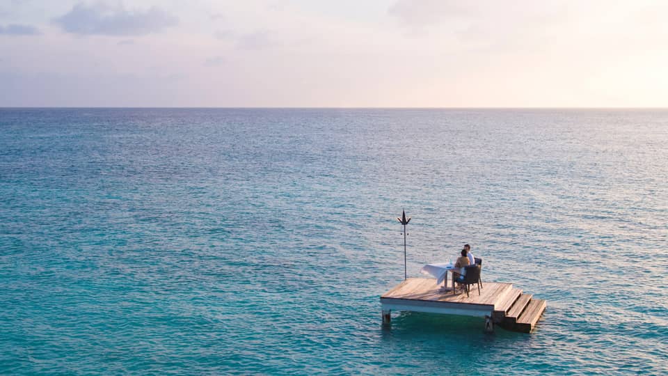 Man, woman dine at private table on small floating wood platform in middle of blue lagoon