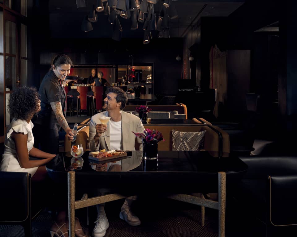 Two people wearing light-coloured cocktail attire sit on benches around a black table as a waitress brings them beverages