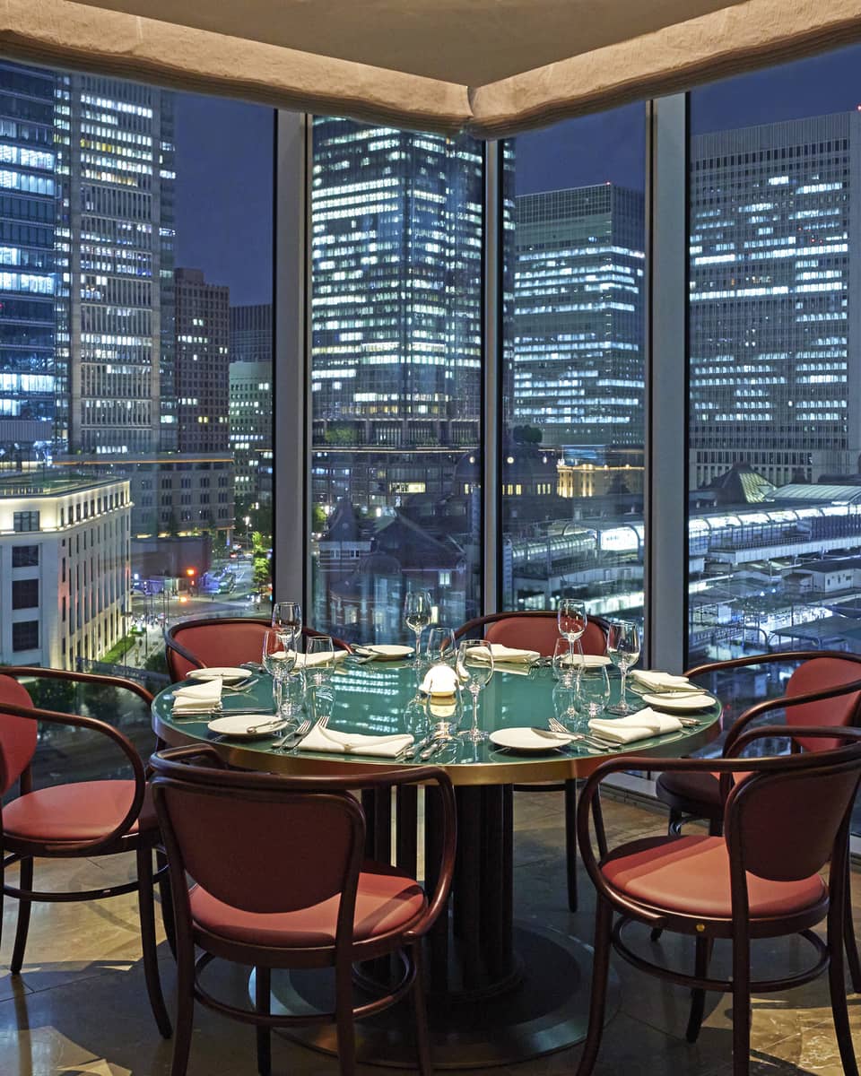 Luxury boutique hotel dining setup with a round table elegantly set for guests, offering a panoramic city view at night through tall glass windows