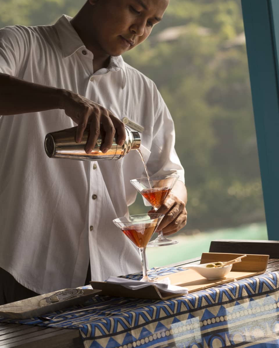 Man in white uniform with silver martini shaker pours pink cocktails into glasses on wood tray