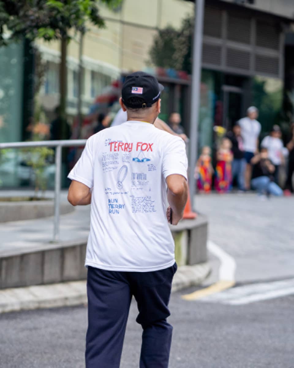 Person in white t-shirt, baseball cap and navy blue pants runs toward crowd of people under balloon arch