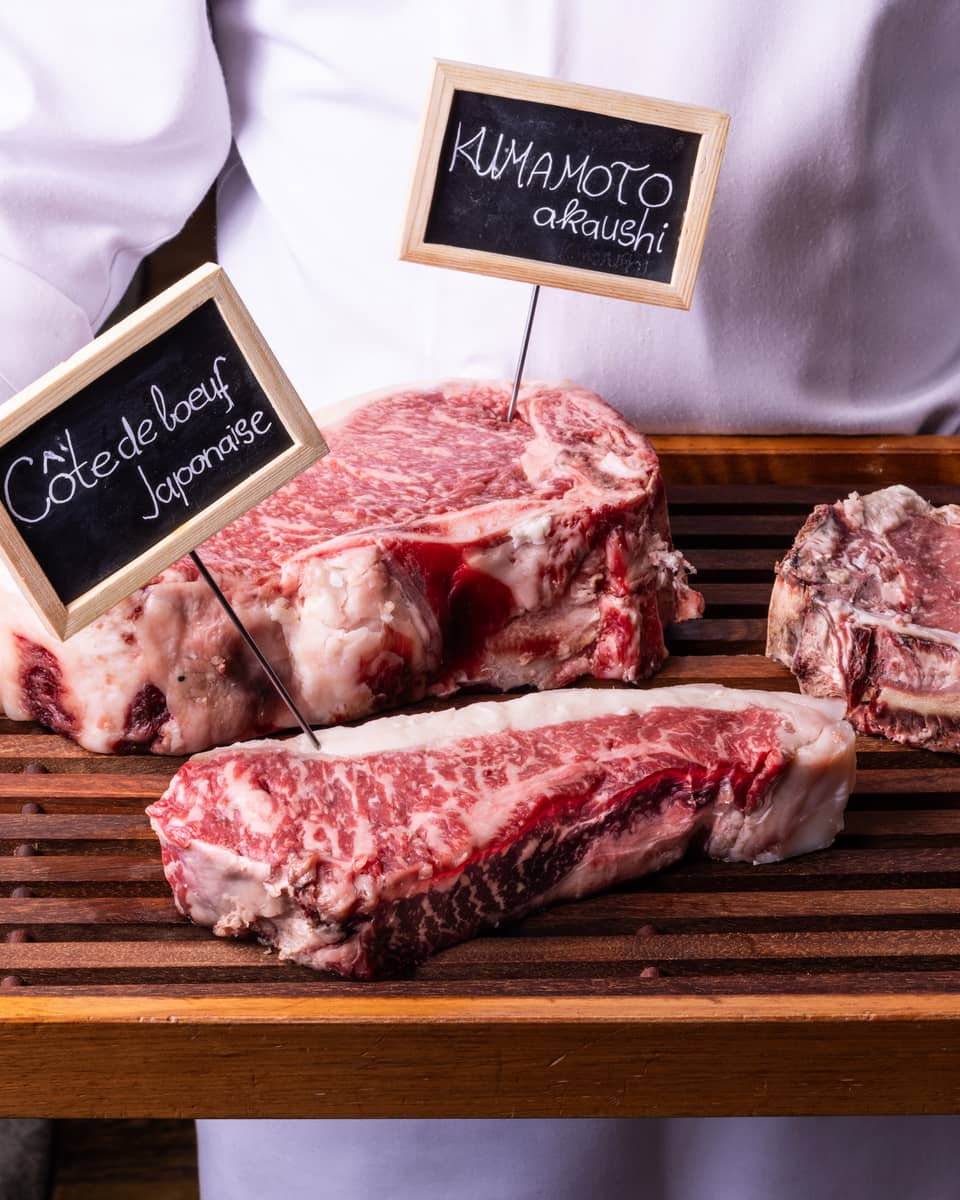 Close-up view of a wood tray with three labelled cuts of well-marbled beef, held by person in white chef's coat.