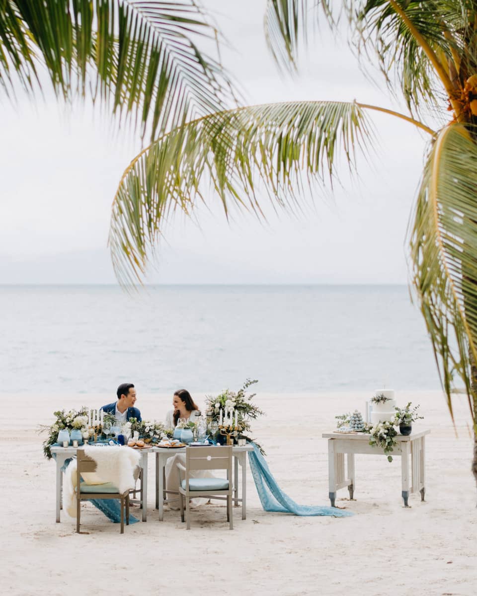 Smiling couple at private dining on white sand beach past palm leaves