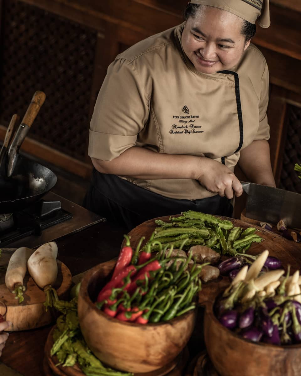 A chef teaching two participants in a cooking class, surrounded by fresh vegetables and ingredients in a rustic kitchen setting.