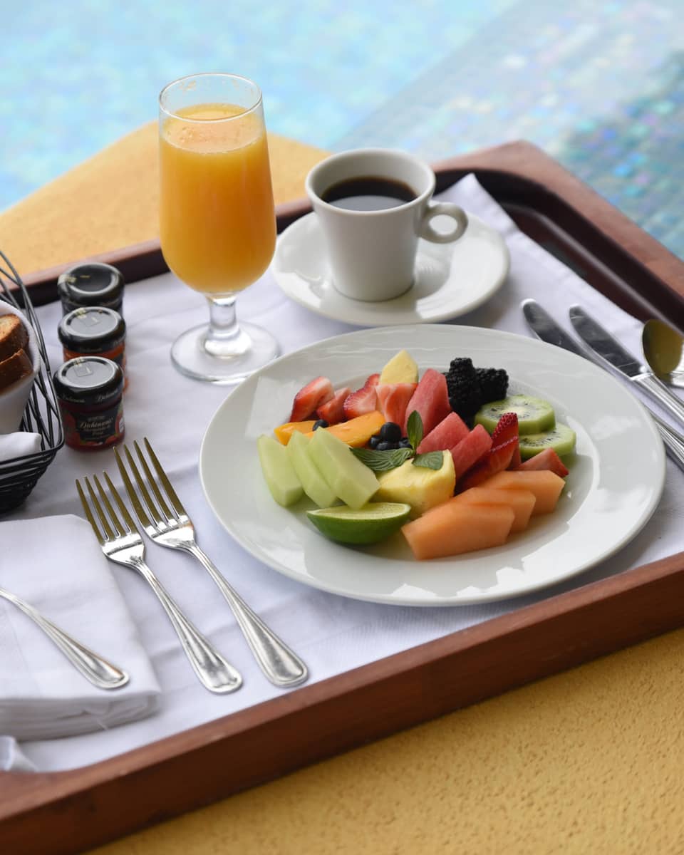 Fresh fruit slices on plate, coffee, orange juice, toast on wood tray by pool