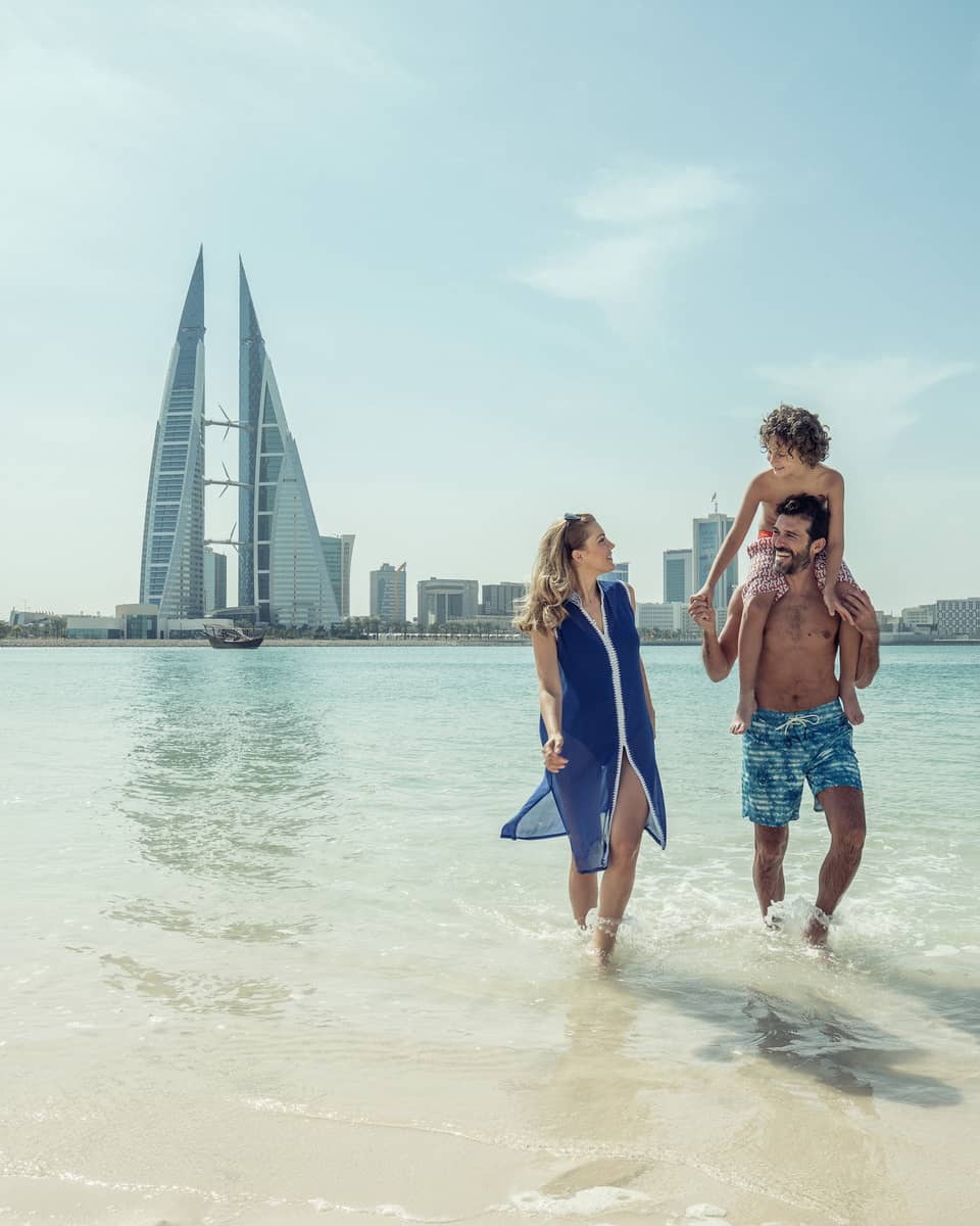 A man with a teenager on his shoulders walks with a woman through shallow ocean water with the city of Bahrain Bay in the background
