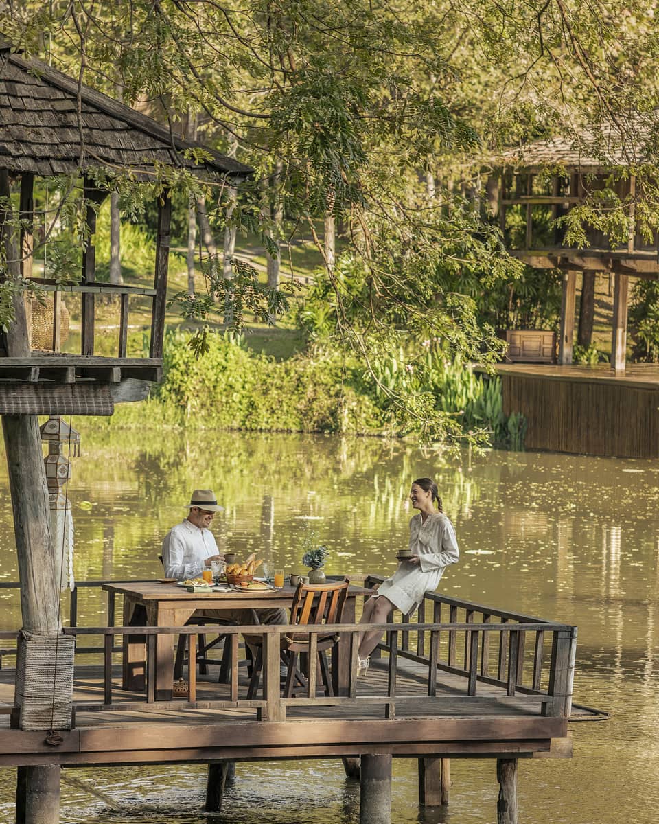 Two guests at a rustic table, on a platform in one of two wood huts on stilts perched atop a calm river bordered by greenery.