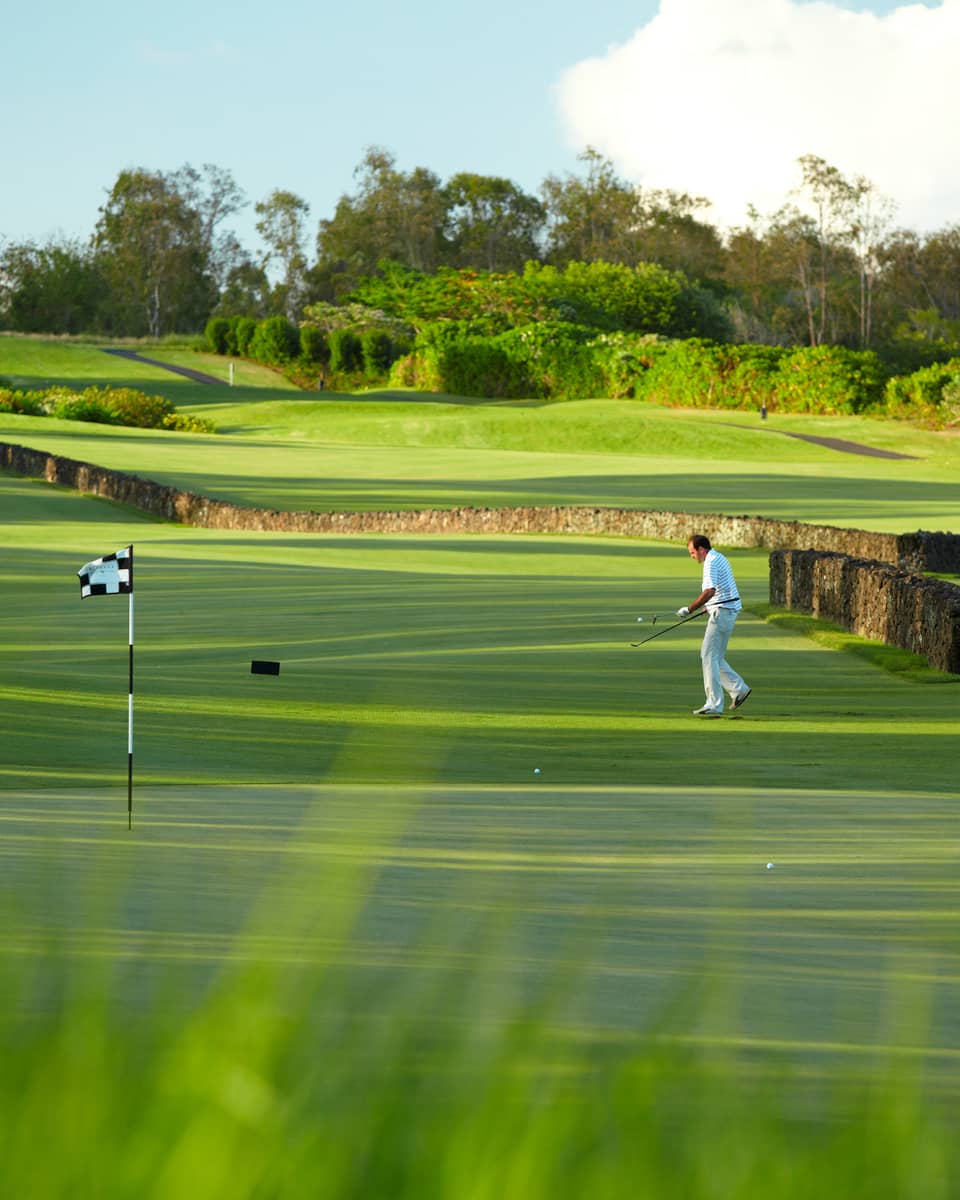 Man with golf club walks toward flag on golf course green near stone fence