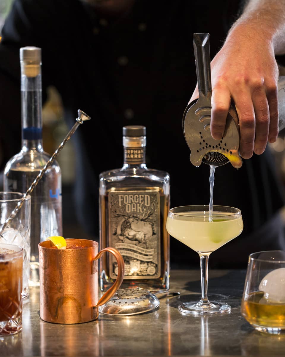 Various cocktail glasses and liquor bottles on bar as mixologist pours martini out of shaker