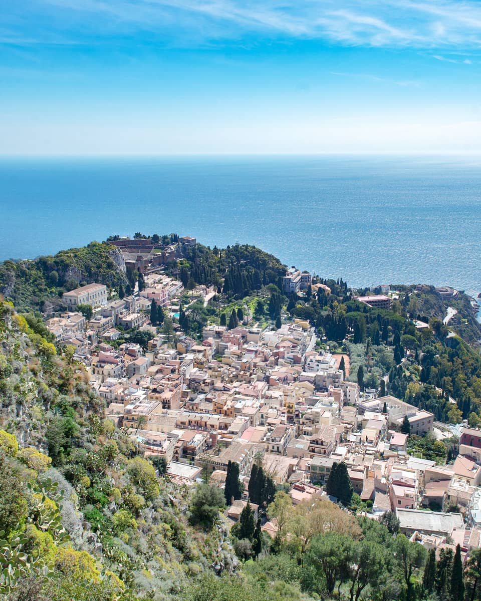 Aerial view of a coastal town with clustered buildings, lush greenery and the blue expanse of the ocean in the background.