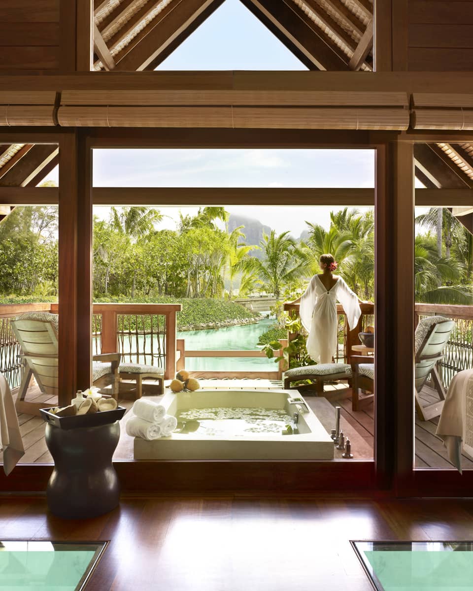 Woman wearing flowing white robe stands at edge of spa suite near plunge bath, couples massage beds