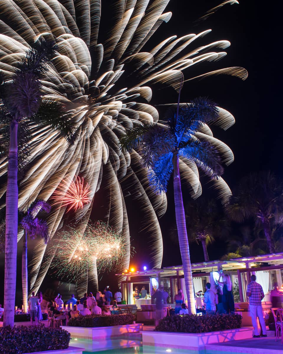 People around resort outdoor pool/bar watching fireworks in the night sky