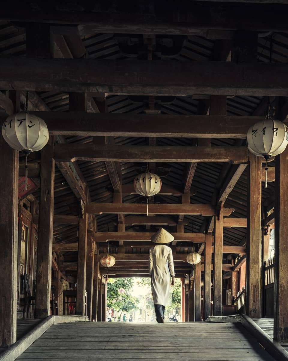 A person wearing traditional Vietnamese attire and a conical hat walks through an ancient wooden outdoor hallway decorated with lanterns.
