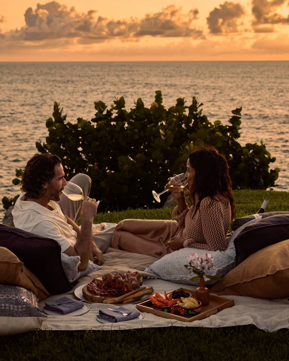 A man and woman lay on blanket and pillows during luxury picnic at sunset, next to the ocean