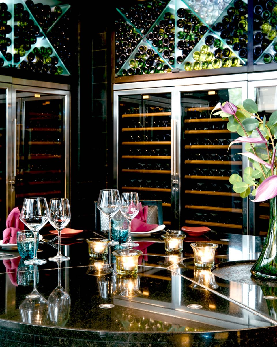 Opulent setting of goblets, candles and a tropical flower atop a mirrored table, surrounded by floor-to-ceiling wine racks.