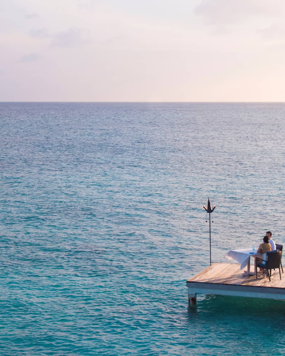 Man, woman dine at private table on small floating wood platform in middle of blue lagoon