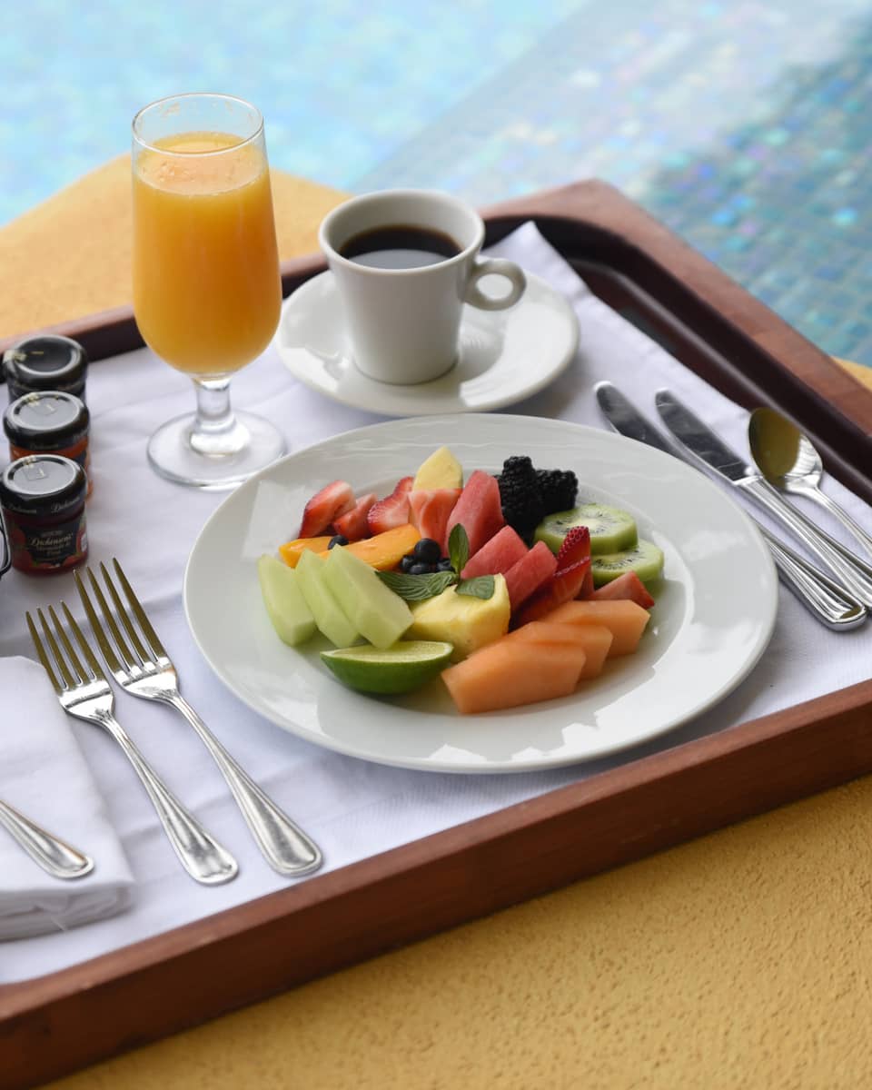 Fresh fruit slices on plate, coffee, orange juice, toast on wood tray by pool