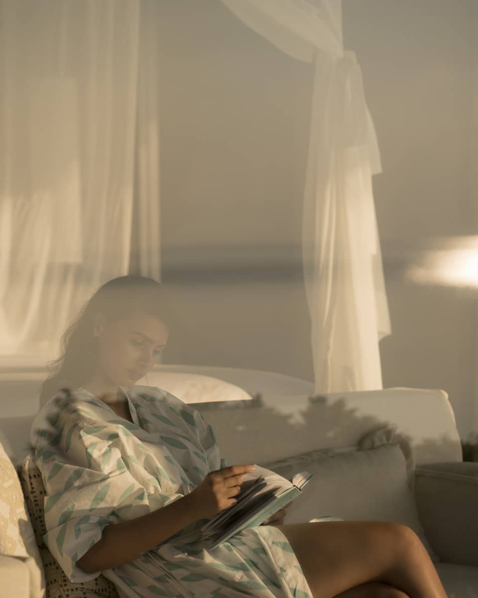 Woman in white robe with green print behind glass, reads book on white sofa