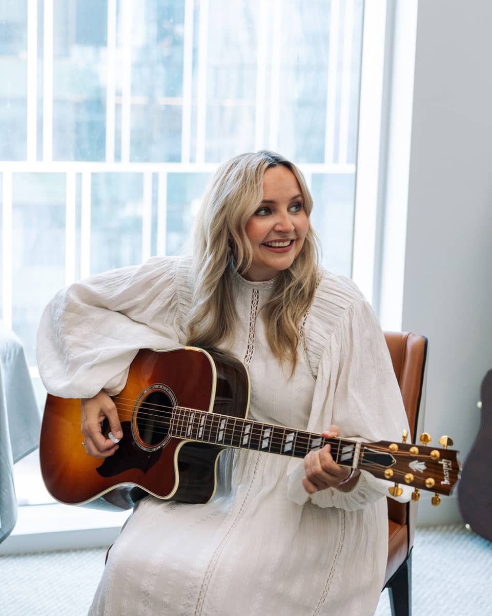 A woman with long blonde hair in a white dress plays an acoustic guitar while seated in a modern room with large windows, smiling and looking to the side.,,A woman with long blonde hair in a white dress plays an acoustic guitar while seated in a modern room with large windows, smiling and looking to the side.